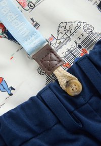 Bretelle bleu clair avec détail en cuir marron fixée à un pantalon bleu marine avec un bouton beige, sur un tissu imprimé coloré à thème londonien.