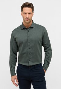 Camicia botton-up verde scuro con tessuto texture, maniche lunghe, colletto a punta e bottoni neri, abbinata a pantaloni blu navy.