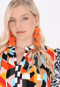Többszínű blúz fodros gallérral, absztrakt mintákkal narancssárgában, feketében, rózsaszínben és kékkel; narancssárga virág fülbevalókkal párosítva.