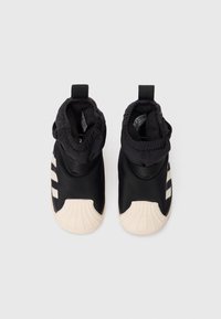 Chaussures pour tout-petits noires et blanches à enfiler avec des poignets élastiques aux chevilles et des embouts en caoutchouc, posées à plat sur un fond blanc.