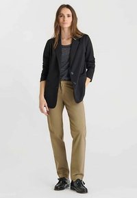 Blazer noir à deux boutons, chemise rayée grise en dessous, pantalon beige et chaussures noires brillantes. Le modèle se tient les mains le long du corps.