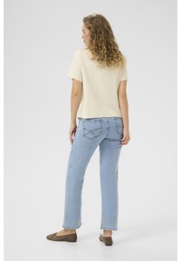 Top beige a maniche corte, vestibilità comoda, indossato con jeans dritti azzurri. Cuciture evidenti sui jeans e ballerine marroni. Sfondo neutro.