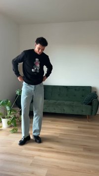 Sweatshirt negru cu un grafic colorat de urs, blugi albastru deschis și încălțăminte neagră; stând pe podea de lemn lângă o canapea verde și o plantă.