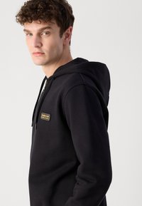 Black zip-up hoodie made from a soft fabric. Features a prominent front logo patch and adjustable drawstring hood. Smooth texture.