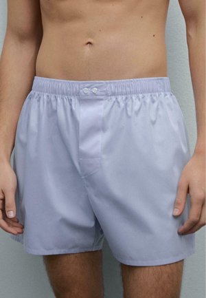 Mężczyzna ubrany w jasnoszare bokserki z elastycznym pasem i guzikiem na przodzie, stojący na tle jednolitego jasnego tła.