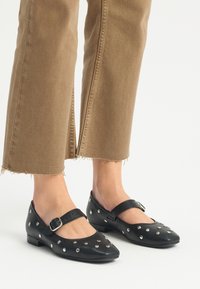 Zapatos Mary Jane de cuero negro con tacón plano, que presentan remaches decorativos plateados, puntera redondeada y una correa ajustable con hebilla.