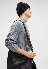 Γκρι ριγωτό πουλόβερ με ψηλό λαιμό, φορεμένο με μαύρο σκουφάκι και κρατώντας μια σκούρα, υφασμάτινη τσάντα ώμου. Πλάγια όψη.