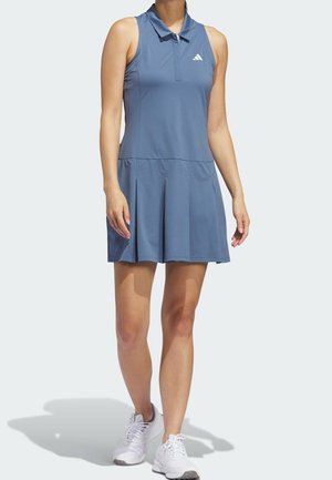 Femme portant une robe de tennis bleue sans manches avec col et chaussures de sport blanches, debout devant un fond uni.