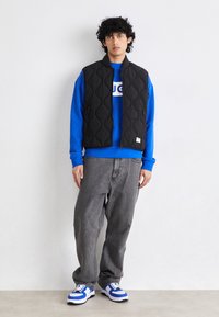 Schwarze gesteppte Weste mit Reißverschluss, blauer Sweatshirt mit weißem Logo, graue Jeans und blau-weiße Sneakers. Schlichtes, lässiges Design.