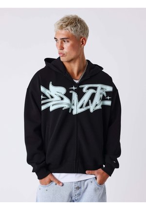 Joven con cabello decolorado que lleva una sudadera negra con cremallera y texto en estilo grafiti azul, pantalones vaqueros claros y un collar de cadena plateada.