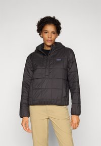 Zwarte gewatteerde jas met een capuchon, voorzien van een kwartrits aan de voorkant en een logopatch. Textuurstof met een gewatteerd ontwerp, gecombineerd met beige broeken.