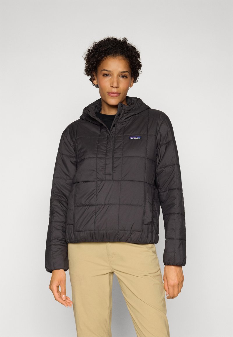 Zwarte gewatteerde jas met een capuchon, voorzien van een kwartrits aan de voorkant en een logopatch. Textuurstof met een gewatteerd ontwerp, gecombineerd met beige broeken.