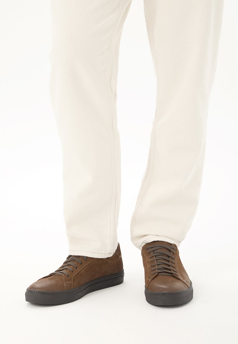 Baskets en cuir marron avec semelles noires portées avec un pantalon clair sur une personne debout sur fond blanc.