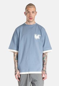 Blå oversized t-shirt med hvid kant ved ærmer og krave, med et lille hvidt logo på brystet. Blødt bomuldsstof.