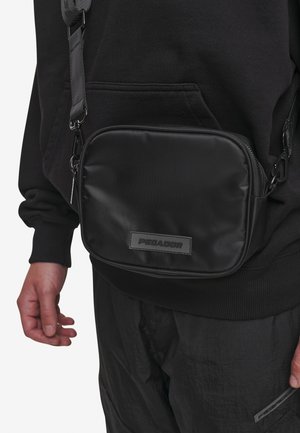 Black nylon crossbody bag with a rectangular shape, zip closure, and a branded patch. Features a sturdy adjustable strap and sleek texture.