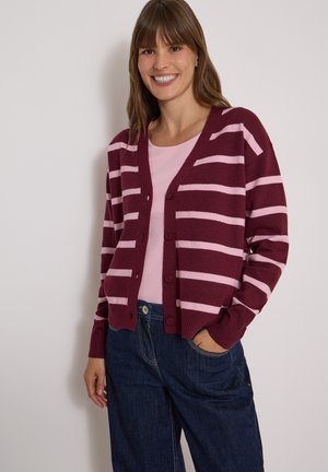 Femme souriante portant un cardigan rayé bordeaux et rose boutonné par-dessus un top rose clair et un jean bleu foncé, debout, une main dans la poche.