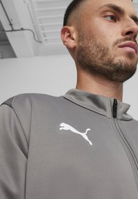 Graue Zip-Jacke aus glattem Material mit hohem Kragen und einem weißen Puma-Logo auf der linken Seite. Nahaufnahme von unten.