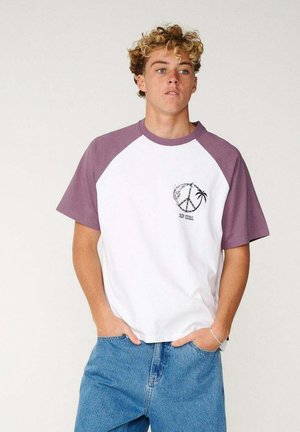 Jeune homme aux cheveux blonds bouclés porte une chemise à manches courtes blanche et violette avec un motif de paix et de palmier et un jean bleu, les mains dans les poches.