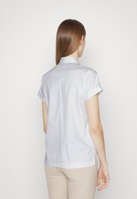 Mujer con cabello castaño lacio, vestida con una camisa blanca de manga corta y pantalones beiges, de espaldas contra un fondo sencillo.