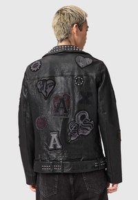 Geacă de motociclist din piele neagră, cu diverse embleme și ținte. Detalii notabile includ modele texturate pe material și o curea decorativă.