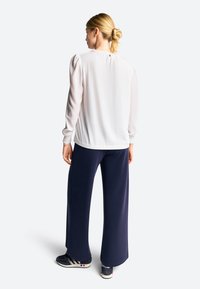 Hvid langærmet bluse med puffede skuldre, lukning med knapper bagpå; sammen med marineblå vide bukser og sorte sneakers.