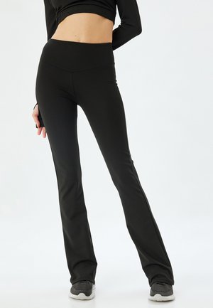 Leggings noires ajustées en matériau extensible, dotées d'une taille haute, d'un bas évasé et d'une texture lisse, accompagnées de chaussures grises.