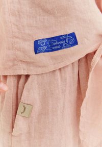 Ružová ľanová látka obsahuje modrú nálepku s nápisom "Saltwater Sunsets" a námornými grafikami, vedľa malej béžovej etikety s dizajnom rias.