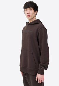 Bruine hoodie gemaakt van zacht materiaal met een voorzak en een gedrukte logo over de borst. Eenvoudig ontwerp met ribgebreide boorden.