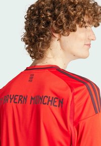 Camiseta de fútbol roja con acentos negros, con "FC BAYERN MÜNCHEN" impreso en la parte trasera y franjas a lo largo de los hombros. Textura suave.