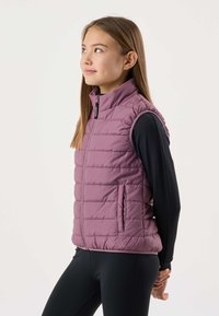 Mlada oseba v črni majici z dolgimi rokavi, oblečena v vijoličasto brezrokavnik, stoji z rokami za hrbtom, gleda navzgor in vstran.