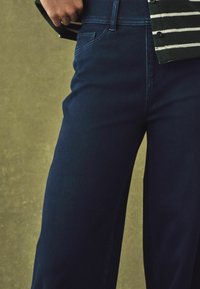 Tumedat sinised denim püksid sileda tekstuuriga. Omavad traditsioonilisi taskuid ja hästi istuvat vöökohta, kombineerituna triibulise nööpidega särgiga.
