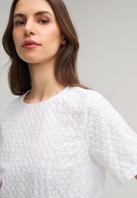 Witte korte mouwen top van textuurstof met een bloemenpatroon. Ronde hals en een ontspannen pasvorm. Subtiele stikdetails zichtbaar.