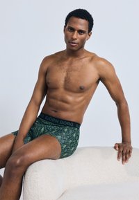 Zelené boxerky s vzorem a černým pasem s názvem značky "BJÖRN BORG." Látka vypadá hladká s jemným leskem.