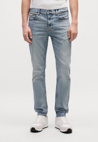 Jeans de mezclilla azul claro con un corte ajustado, que presentan un detalle desgastado en la rodilla derecha, diseñados para usar en ocasiones casuales. Combinados con zapatillas blancas.