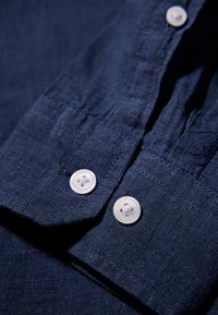 Donkerblauw, gestructureerd stoffen shirt met een mouw waar twee witte knopen vastgemaakt zijn aan de manchette, met gedetailleerde stitching.