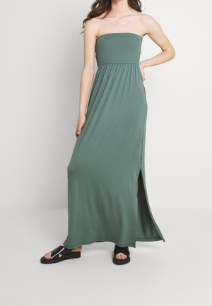 Femme portant une robe maxi olive verte sans bretelles avec une fente latérale et des sandales à plateforme noires à bouts ouverts, debout devant un fond uni.