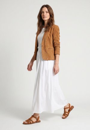 Vrouw loopt, draagt een witte jurk, een beige suède jas en bijpassende bruine sandalen met gesp tegen een egale lichte achtergrond.