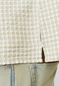Camisa texturizada en beige claro con un patrón geométrico, que presenta una abertura lateral y una etiqueta. Combinada con unos jeans de mezclilla azul claro.