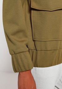 Olivgrön jacka i ett texturerat tyg, med en vikbar ficka och elastiska ärmslut. Synliga sömmar ger extra djup åt designen.