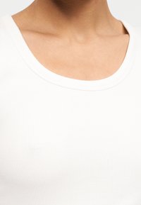 Λαιμός και άνω μέρος του στήθους γυναίκας με λευκό ριμπ μπλουζάκι με λαιμόκοψη, εμφανιζόμενα πάνω σε απλό λευκό φόντο.