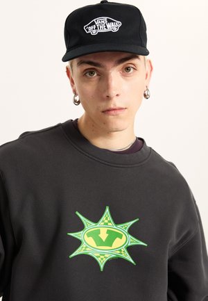 Felpa nera con una grande grafica verde e gialla a forma di esplosione stellare. Chi la indossa porta anche un cappellino nero con un logo bianco.