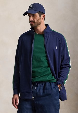 Férfi, aki tengerészkék sapkát, tengerészkék cipzáras dzsekit zöld csíkokkal, zöld inget, tengerészkék nadrágot és karórát visel, oldalra néz, az egyik kezét a zsebében tartva.