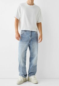 Vit kortärmad t-shirt i kombination med ljusblå denimjeans som har ett präglat mönster. Avslutat med krämfärgade sneakers.