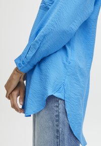 Camicia azzurra chiara con texture, a maniche lunghe, caratterizzata da un orlo curvo e polsini con bottoni, abbinata a jeans chiari in denim.