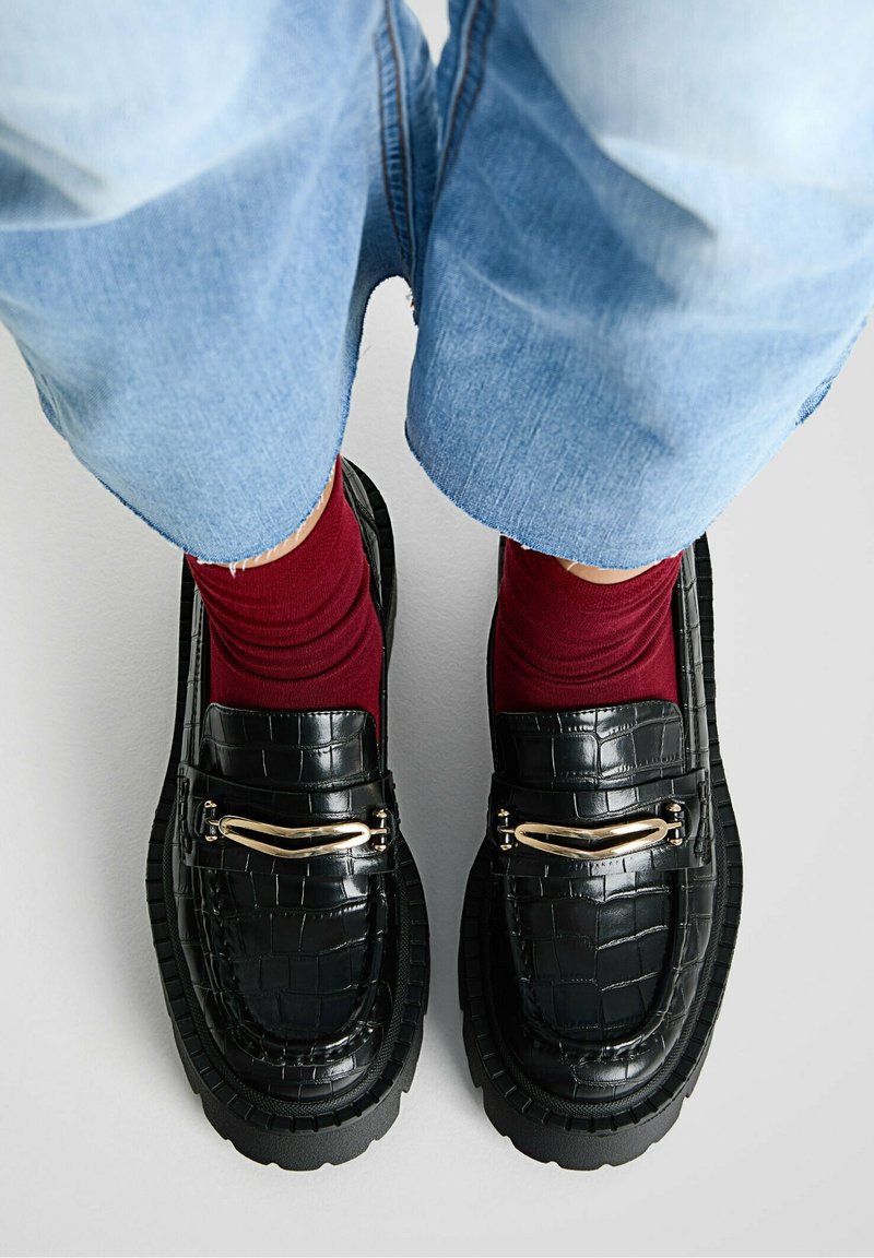 Mocassins en cuir synthétique noir effet crocodile avec ferrures dorées, semelle en caoutchouc noire épaisse, portés avec des chaussettes bordeaux et un pantalon en denim bleu retroussé.