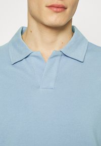 Dolní část obličeje a krk muže, který nosí světle modrou polokošili s límečkem, na jednobarevném pozadí.