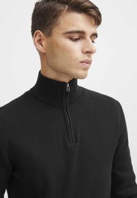 Schwarzer Zip-Kragen-Pullover mit geripptem Kragen, gestricktem Textil und eng anliegendem Design, das einen kleinen Reißverschluss am Hals hat.