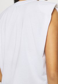 Biała bezrękawnikowa bluzka o luźnym kroju, z lekko przedłużonymi liniami ramion i miękkim bawełnianym materiałem, prezentująca gładką teksturę.