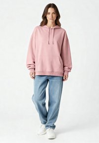 Felpa oversize rosa con coulisse, tasca frontale e orlo a coste, abbinata a jeans azzurri e sneakers bianche. Tessuto morbido.