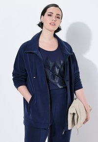 Tuta in velluto blu navy con giacca con zip, vestibilità ampia e pantaloni a gamba stretta. La parte superiore presenta un design grafico floreale nero. Tiene in mano una pochette beige.